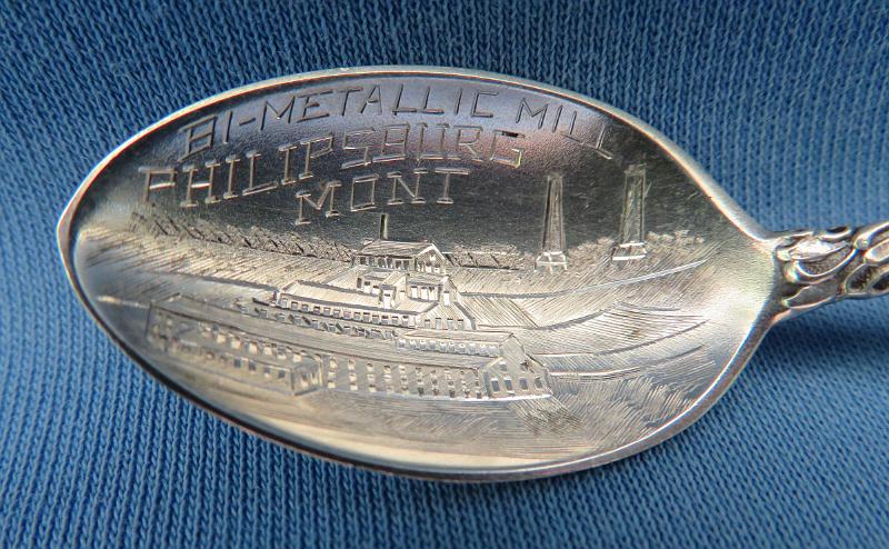 Souvenir Mining Spoon Bi-Metallic Mill Bowl.JPG - SOUVENIR MINING SPOON BI-METALLIC MILL PHILIPSBURG MT - Sterling silver souvenir demitasse spoon, 4 1/4 in. long, bowl engraved with mill buildings and marked BI-METALLIC MILL PHILIPSBURG MONT., handle marked in floral pattern, reverse marked Sterling with maker’s mark C.F.R.  [The historic mining town of Philipsburg, Montana is located about 55 miles northwest of Butte.  Founded in 1867 and named for Philip Deidesheimer of Comstock fame, the area around Philipsburg was home to the Granite Mountain and Bi-Metallic mines, major producers of the greatest silver bonanza at the time.  Both mines worked the same ore body first located in 1872.  In 1882 Charles McLure and  partners organized the Bi-Metallic Mining Company.  The company built the Bi-Metallic mill in 1888 along Douglas Creek about one mile southeast of Philipsburg in the small town known as Kirkland and later Clark.  The 50-stamp mill, 150 feet wide and 367 feet long, was rated at 75 tons with the capacity to add more stamps later.  At the time it was the largest stamp mill in Montana.  The town, which reached a peak population of about 125 people, was primarily inhabited by mill workers. It once included numerous cabins, a boarding house, company office, warehouse, and assay office, along with the large Bi-Metallic mill site. The mine and mill employed 500 workers. To get the ore to the mill, a tramway was constructed in 1889 from the mine to the Bi-Metallic mill, 9750 feet long with a vertical drop of 1225 feet, which was the longest aerial tramway in the United States at that time.  The mill was expanded to 100 stamps in 1891 giving it a 200-ton capacity. A huge 4,307 pound bar of silver bullion from the Bi-Metallic was displayed at the 1893 World's Fair.  From 1883 to 1893, the Bi-Metallic produced $6 million in silver and paid dividends of $2 million. Several years after the silver panic of 1893 the ownership of the Granite and Bi-Metallic was consolidated and years of intermittent company and leasing operations followed. . Operations ended in1918 as silver mining in the district gave way to manganese.  In 1967, the 367 feet long mill building was burned for safety reasons, but its two smokestacks and foundations continue to stand.]